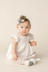 a 9 month old baby girl sitting for photoshoot wearing a white dress